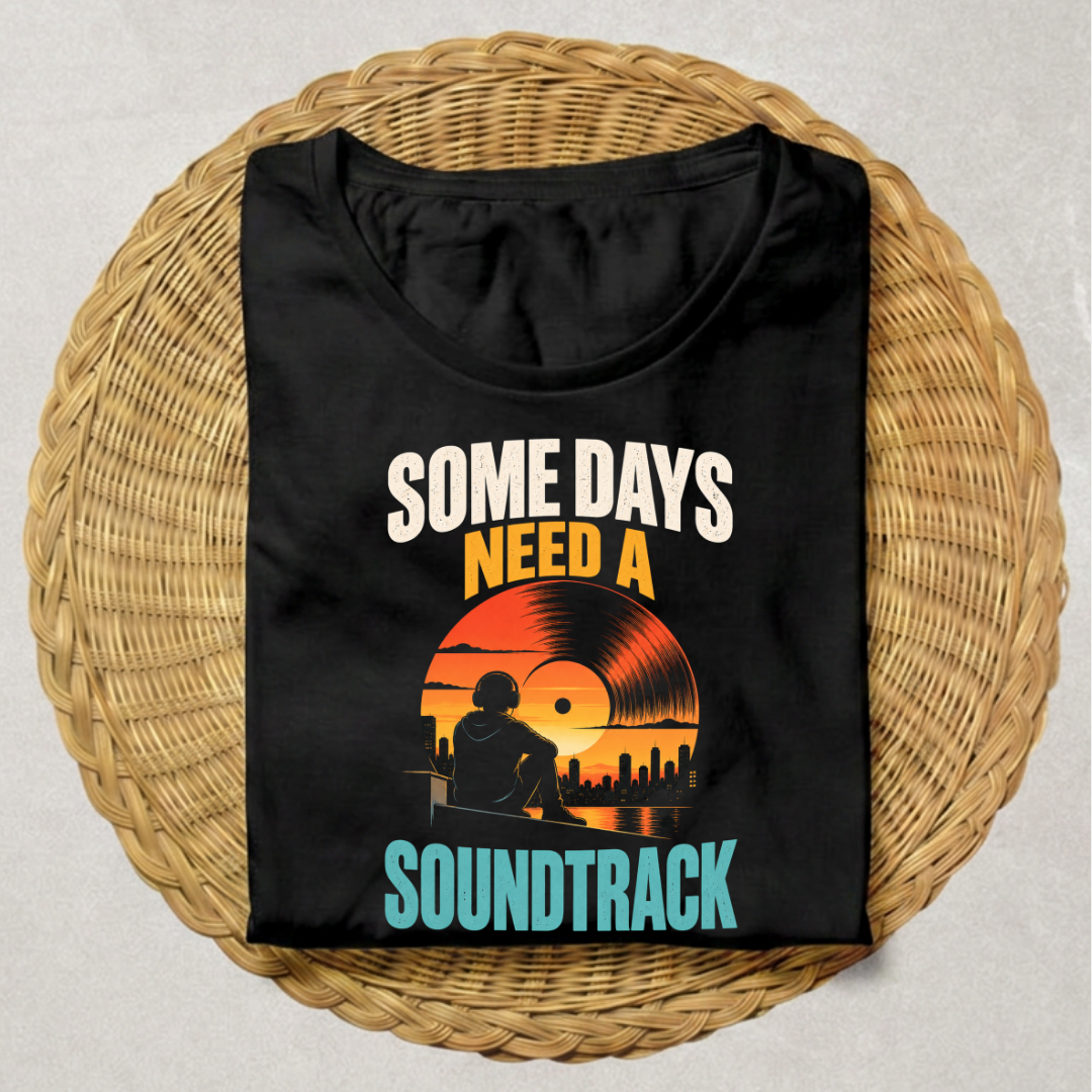 A black short-sleeve t-shirt with a retro-style graphic print reading 'SOME DAYS NEED A SOUNDTRACK' in white, orange, yellow, and teal, displayed flat on a round woven wicker mat against a light gray background.