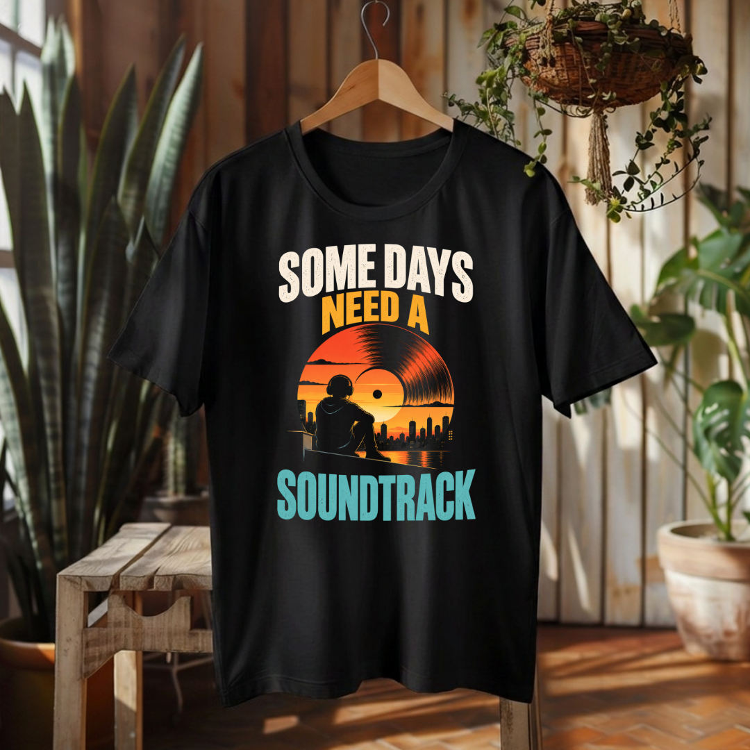 A black short-sleeve graphic T-shirt hanging on a wooden hanger in a rustic indoor setting with plants and wood paneling. The shirt features a retro-style vinyl record graphic with the text 'SOME DAYS NEED A SOUNDTRACK' in white, orange, and teal lettering.