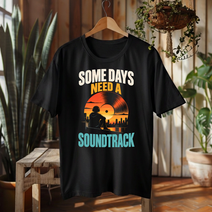 A black short-sleeve graphic T-shirt hanging on a wooden hanger in a rustic indoor setting with plants and wood paneling. The shirt features a retro-style vinyl record graphic with the text 'SOME DAYS NEED A SOUNDTRACK' in white, orange, and teal lettering.