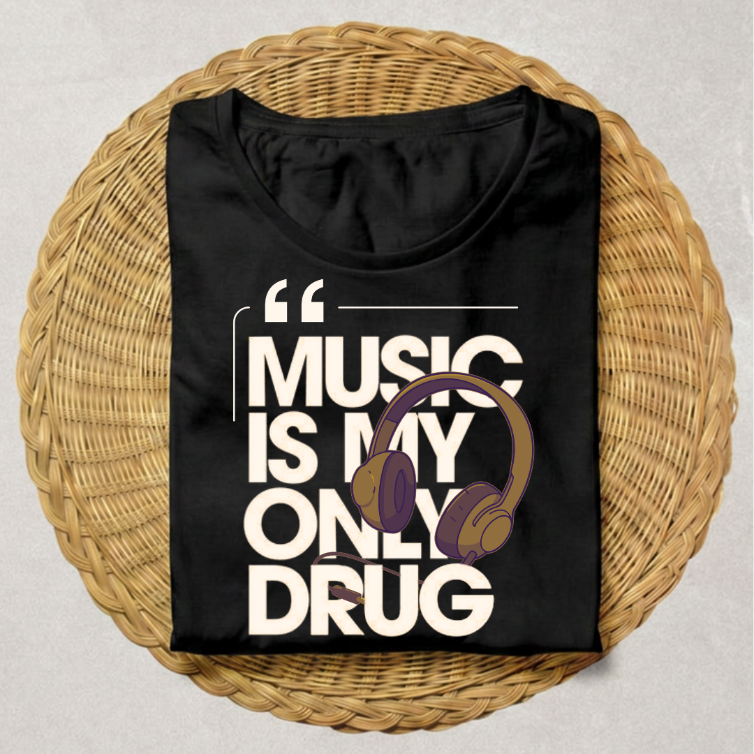A black short-sleeve t-shirt with a graphic print reading 'MUSIC IS MY ONLY DRUG' in bold white letters, featuring a purple and gold headphone illustration. The shirt is neatly folded and presented on a round woven wicker placemat over a neutral gray background.