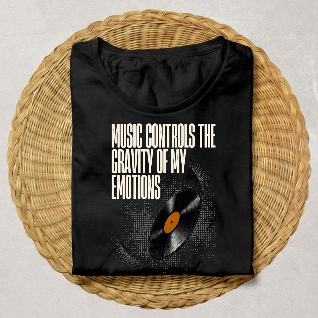 A black short-sleeve T-shirt laid flat on a round woven wicker tray, featuring the white text 'MUSIC CONTROLS THE GRAVITY OF MY EMOTIONS' above a vinyl record graphic with a black-and-white striped pattern and a small orange center label.