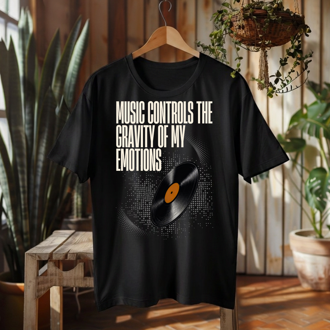 A black graphic t-shirt hanging on a wooden hanger in a rustic indoor setting. The shirt features the text 'MUSIC CONTROLS THE GRAVITY OF MY EMOTIONS' in white, above a graphic of a vinyl record with a black and white pixelated circle and a small orange center label.