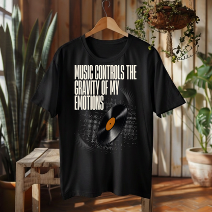 A black graphic t-shirt hanging on a wooden hanger in a rustic indoor setting. The shirt features the text 'MUSIC CONTROLS THE GRAVITY OF MY EMOTIONS' in white, above a graphic of a vinyl record with a black and white pixelated circle and a small orange center label.
