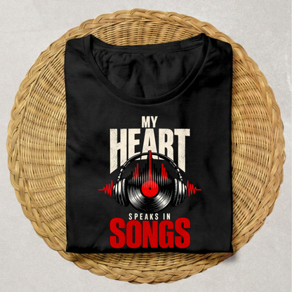 A black short-sleeve t-shirt with a graphic print reading 'MY HEART SPEAKS IN SONGS' in white and red, featuring a vinyl record and headphones design, displayed flat on a round woven wicker mat.