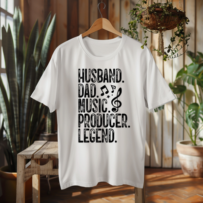 A white short-sleeve T-shirt hanging on a wooden hanger in a rustic indoor setting with plants and wood paneling. The shirt has bold black text reading “HUSBAND.