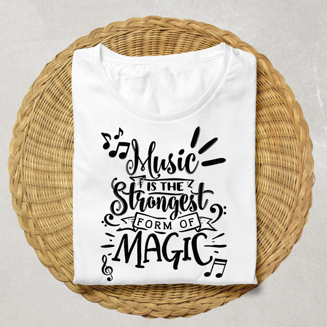 A folded white t-shirt with a black graphic text reading 'Music is the strongest form of magic' and musical notes, displayed on a round woven wicker tray against a light gray background.