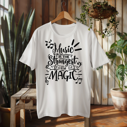 A white short-sleeve graphic T-shirt hanging on a wooden hanger in a rustic indoor setting with plants and wood paneling. The shirt features a black hand-lettered graphic that reads, 'Music is the strongest form of magic' with musical notes and decorative flourishes.