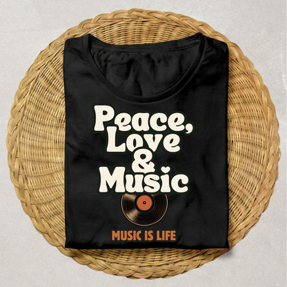 A black short-sleeve T-shirt laid flat on a round woven wicker tray, featuring the phrase 'Peace, Love & Music' in large white retro lettering above a vinyl record graphic, with 'MUSIC IS LIFE' printed below in orange.