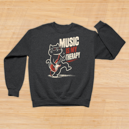A dark heather gray crewneck sweatshirt laid flat on a light wood background. The sweatshirt features a graphic of a cartoon cat playing a red electric guitar, with the text 'MUSIC IS MY THERAPY' printed in white and red.