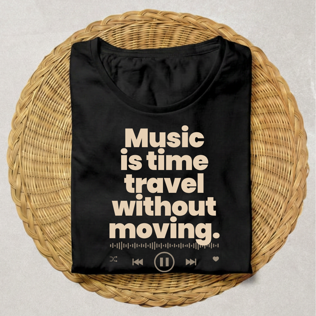 A black short-sleeve T-shirt laid flat on a round woven wicker tray, with large cream text reading 'Music is time travel without moving.' and small music-player interface icons along the bottom.