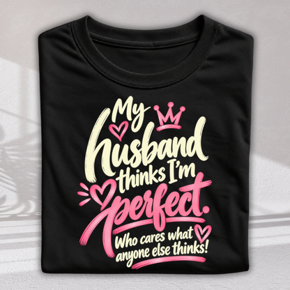 A folded black t-shirt with a humorous graphic print that reads 'My husband thinks I'm perfect. Who cares what anyone else thinks!' in cream and pink lettering with a small crown and heart details, displayed on a light gray background.