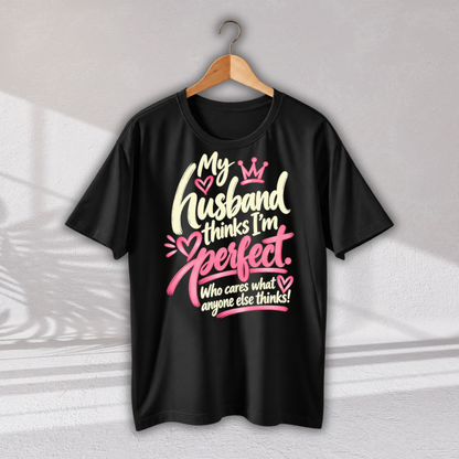 A black short-sleeve t-shirt hanging on a wooden hanger against a light gray wall. The shirt features a graphic print with the text 'My husband thinks I'm perfect.