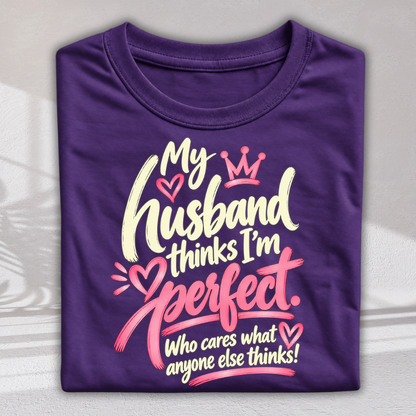 A folded purple t-shirt with a graphic print that reads 'My husband thinks I'm perfect. Who cares what anyone else thinks!' in white and pink lettering with hearts and a crown.
