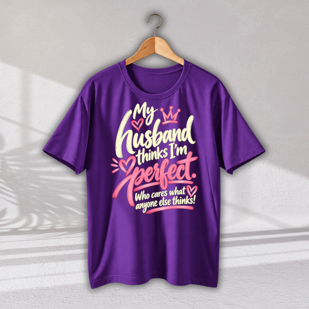 A purple short-sleeve t-shirt hanging on a wooden hanger against a light gray wall. The shirt features a graphic print on the front with the text 'My Husband thinks I'm perfect.