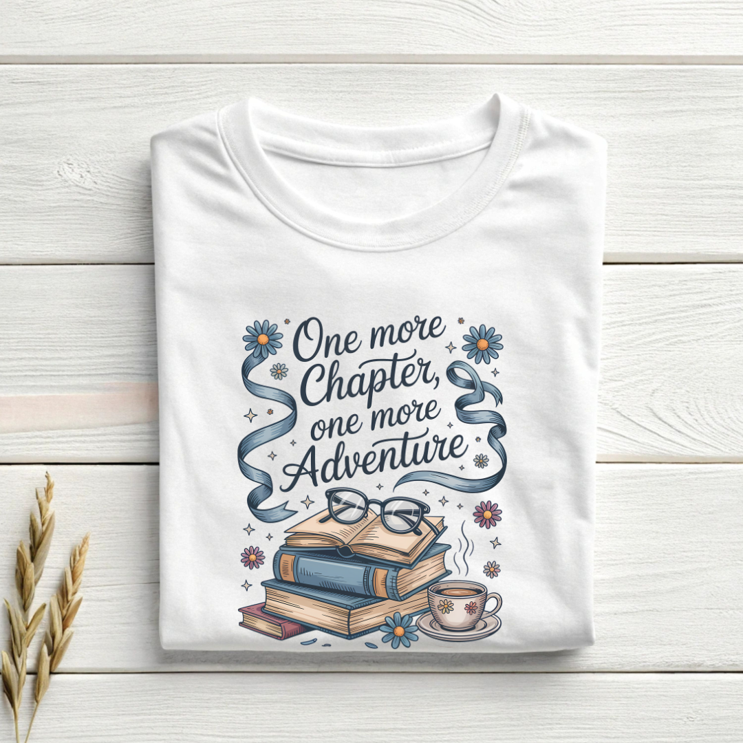 White t-shirt with book-themed design and text 'One more Chapter, one more Adventure' on a wooden surface.