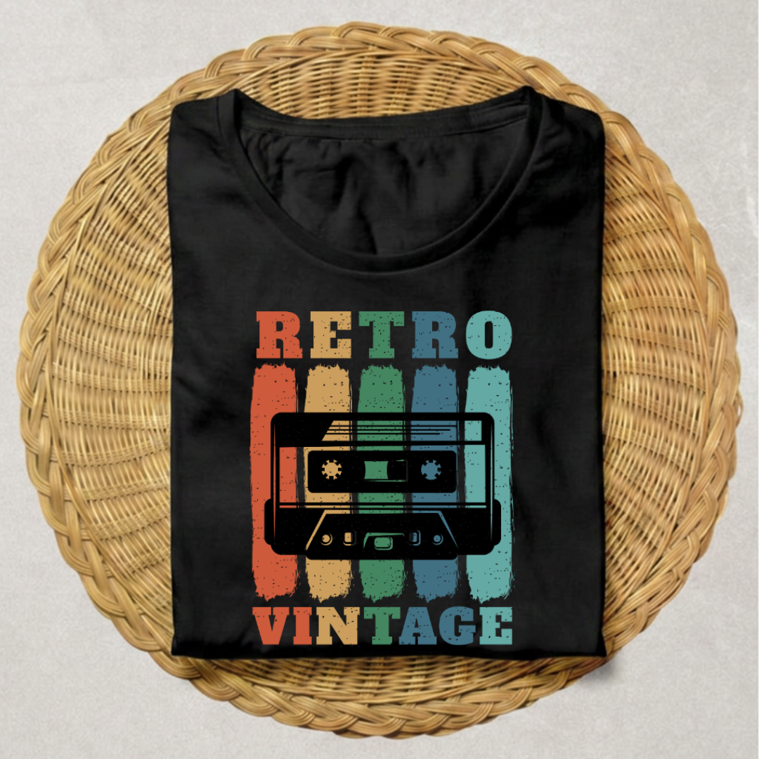A black short-sleeve T-shirt laid flat on a round woven wicker tray, featuring a retro cassette tape graphic with the words 'RETRO VINTAGE' in distressed multicolor lettering.