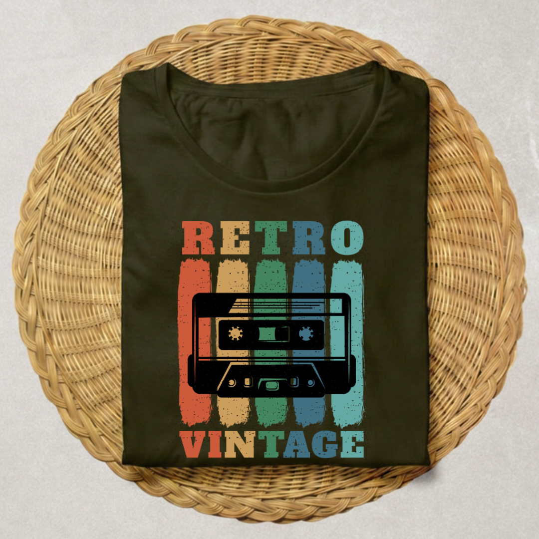 A folded dark olive green t-shirt with a retro cassette tape graphic and the words 'RETRO VINTAGE' in distressed multicolor lettering, displayed on a round woven wicker placemat over a light gray surface.