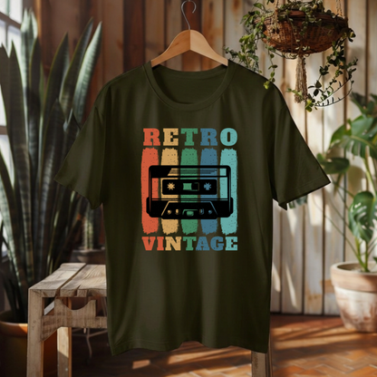 An olive green short-sleeve t-shirt on a wooden hanger, featuring a retro cassette tape graphic with the words 'RETRO VINTAGE' in distressed orange, yellow, teal, and blue colors. The shirt is displayed in a warm, rustic indoor setting with plants and wooden furniture.