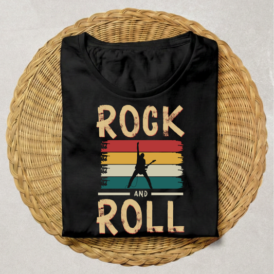 A black short-sleeve t-shirt with a retro graphic reading 'ROCK AND ROLL' in distressed beige letters over a striped sunset design, displayed flat on a round woven wicker tray against a light gray background.