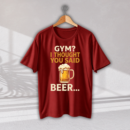 A dark red short-sleeve t-shirt hanging on a wooden hanger against a light gray wall. The shirt features a graphic print with the text 'GYM? I THOUGHT YOU SAID BEER.