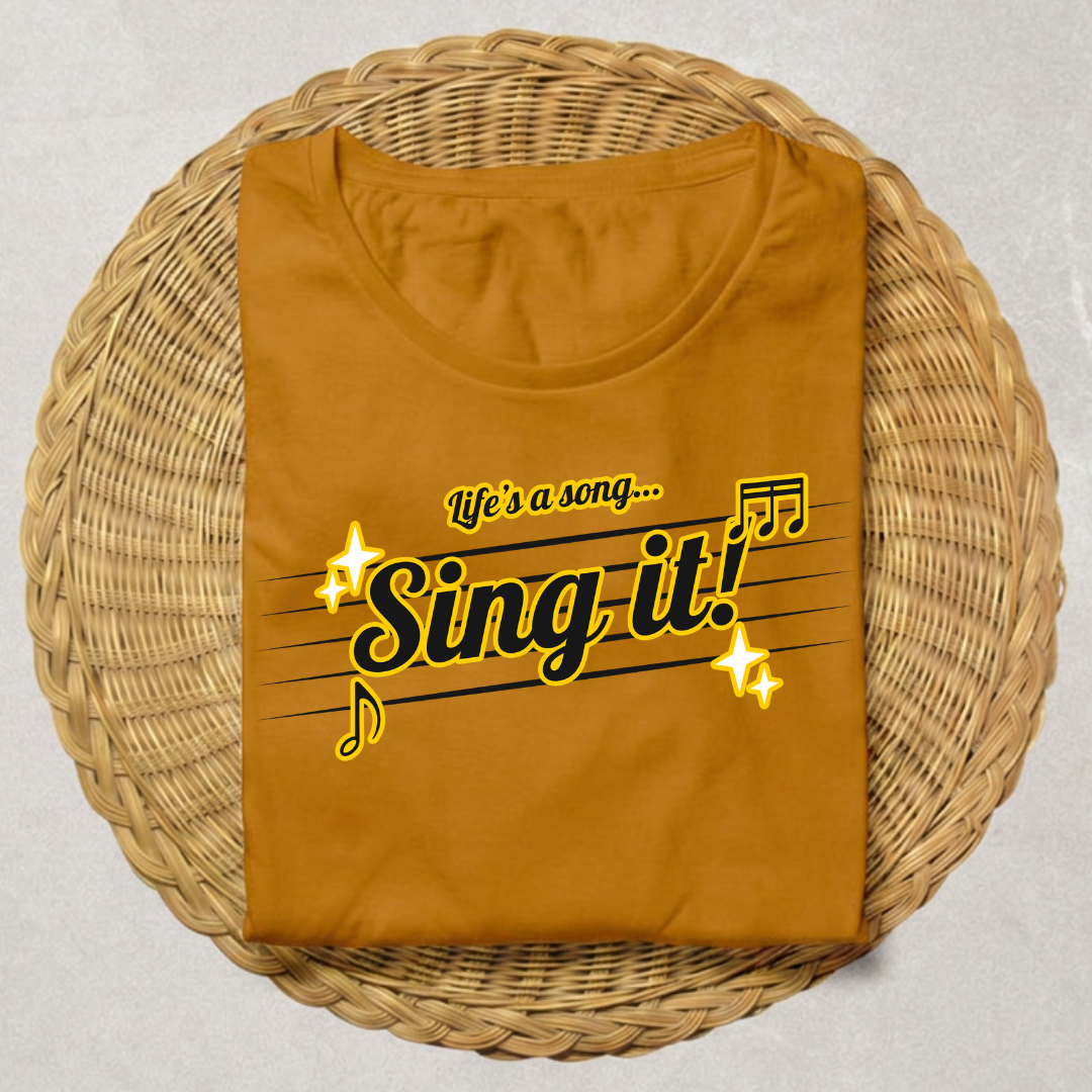 A folded mustard yellow t-shirt with a 'Sing it!' graphic printed on the front, displayed on a round woven wicker tray over a light gray background.