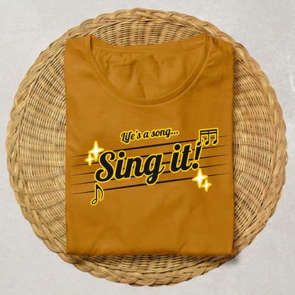 A folded mustard yellow t-shirt with a 'Sing it!' graphic printed on the front, displayed on a round woven wicker tray over a light gray background.
