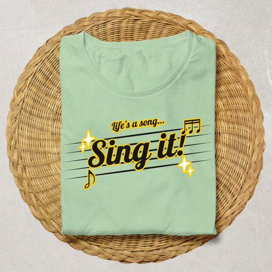 A folded mint green t-shirt with a 'Sing it!' music note graphic, displayed on a round woven wicker tray over a neutral gray background.