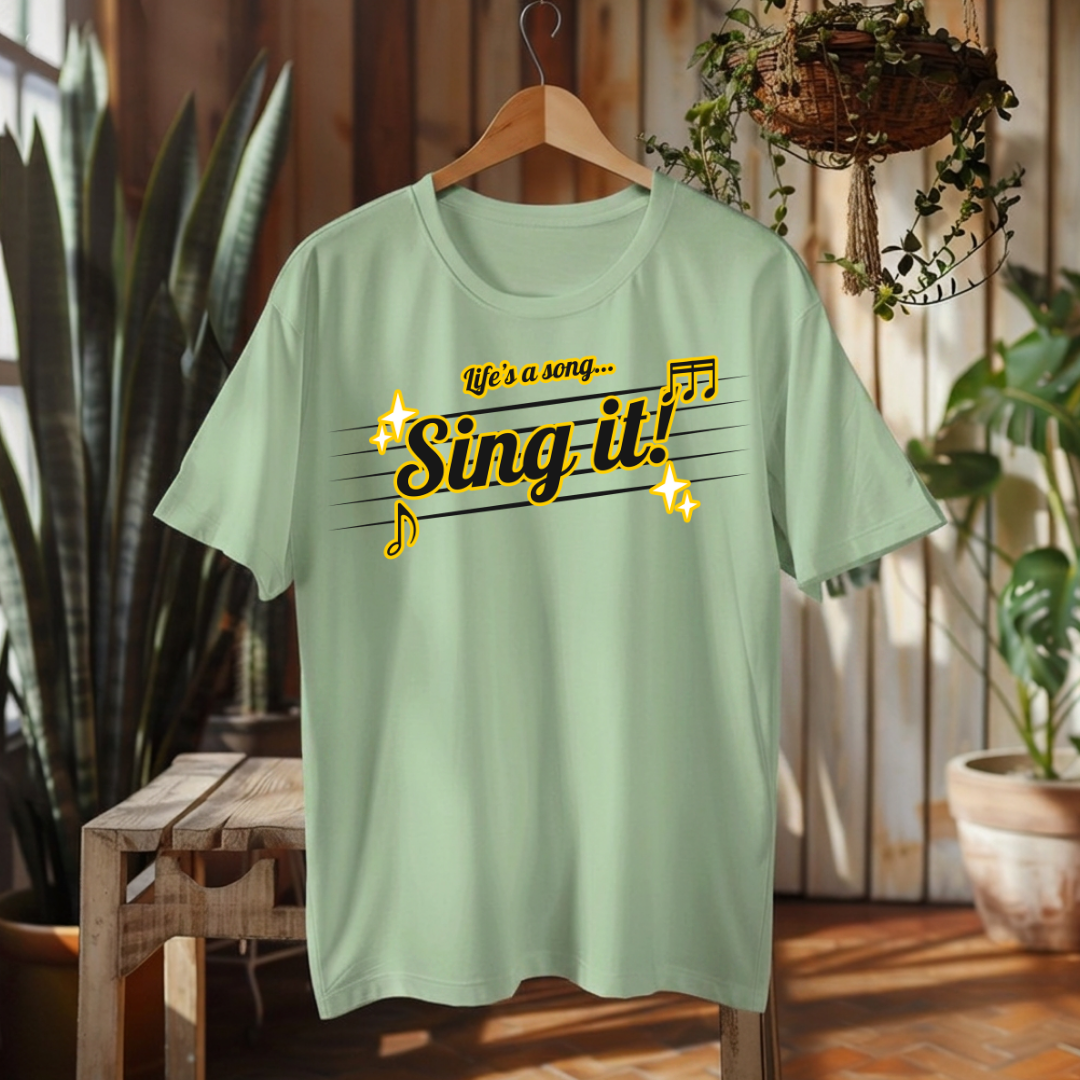 A sage green short-sleeve t-shirt hanging on a wooden hanger in a rustic indoor setting. The shirt features a graphic print on the front with the text 'Life's a song.