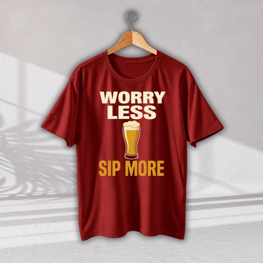 A red short-sleeve t-shirt hanging on a wooden hanger against a light gray wall. The shirt features a graphic print with the text 'WORRY LESS SIP MORE' in cream and yellow, with a pint of beer in the center.