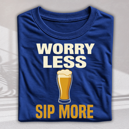 A folded royal blue t-shirt with a graphic print that reads 'WORRY LESS SIP MORE' in white and yellow text, featuring an illustration of a pint of beer in the center, displayed on a light gray textured background.