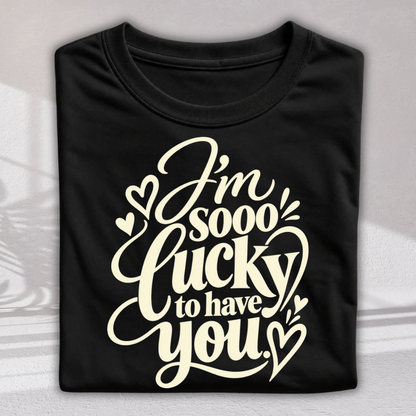 A folded black t-shirt with a cream-colored graphic text that reads 'I'm sooo lucky to have you' decorated with small hearts, displayed on a light gray background.