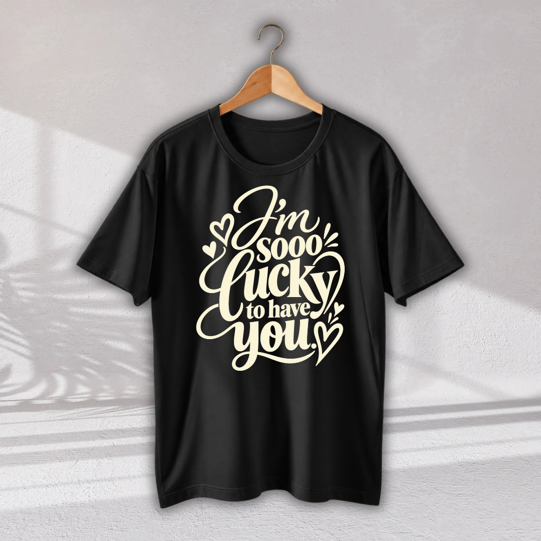 A black short-sleeve t-shirt hanging on a wooden hanger against a light gray wall. The shirt features a large cream-colored graphic text that reads 'I'm sooo lucky to have you' decorated with small hearts.