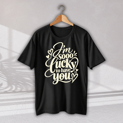 A black short-sleeve t-shirt hanging on a wooden hanger against a light gray wall. The shirt features a large cream-colored graphic text that reads 'I'm sooo lucky to have you' decorated with small hearts.