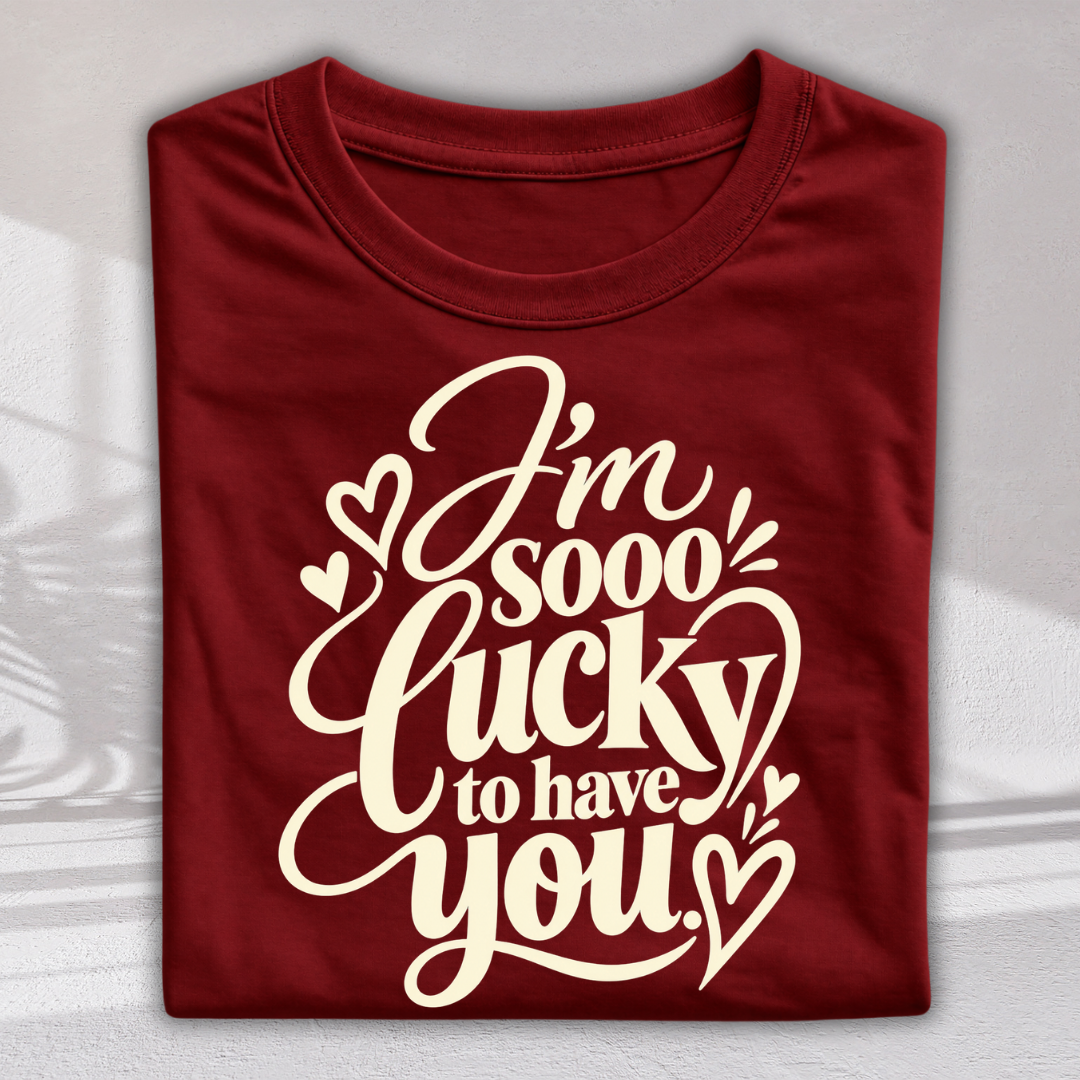 A folded maroon crewneck t-shirt with a cream-colored graphic text that reads 'I'm sooo lucky to have you' surrounded by small hearts, displayed on a light gray background with soft shadows.