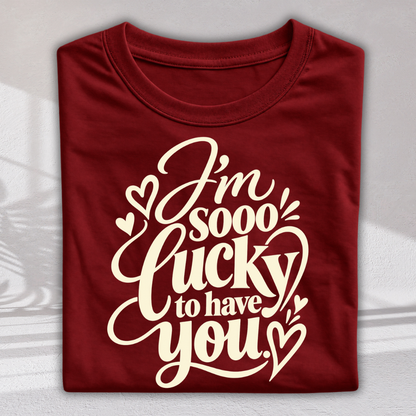 A folded maroon crewneck t-shirt with a cream-colored graphic text that reads 'I'm sooo lucky to have you' surrounded by small hearts, displayed on a light gray background with soft shadows.