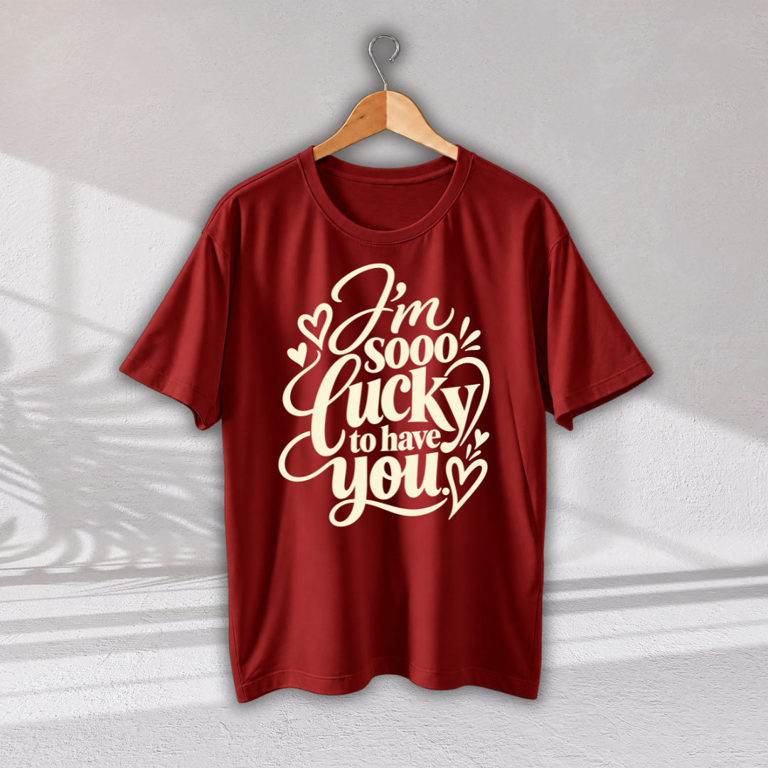 A red short-sleeve t-shirt hanging on a wooden hanger against a light gray wall. The shirt features a large white graphic text on the front that reads 'I'm sooo lucky to have you' decorated with small hearts.