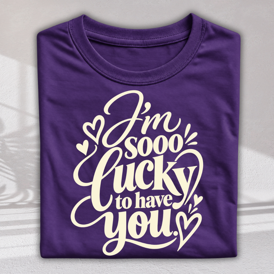 A folded purple t-shirt with a white graphic print that reads 'I'm sooo lucky to have you' decorated with small hearts, shown on a light gray background with soft shadow overlays.