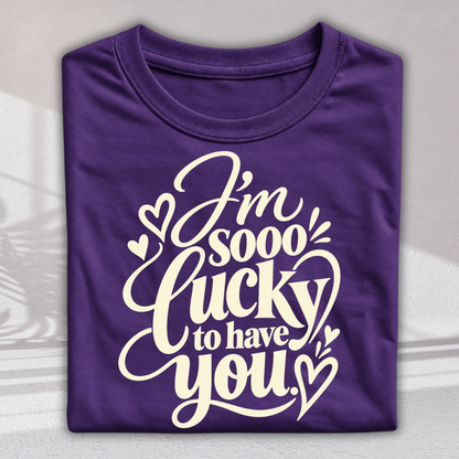 A folded purple t-shirt with a white graphic print that reads 'I'm sooo lucky to have you' decorated with small hearts, shown on a light gray background with soft shadow overlays.
