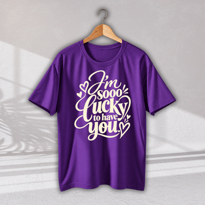 A purple short-sleeve t-shirt hanging on a wooden hanger against a light gray wall. The shirt features a white graphic text design that reads 'I'm sooo lucky to have you' decorated with small hearts.