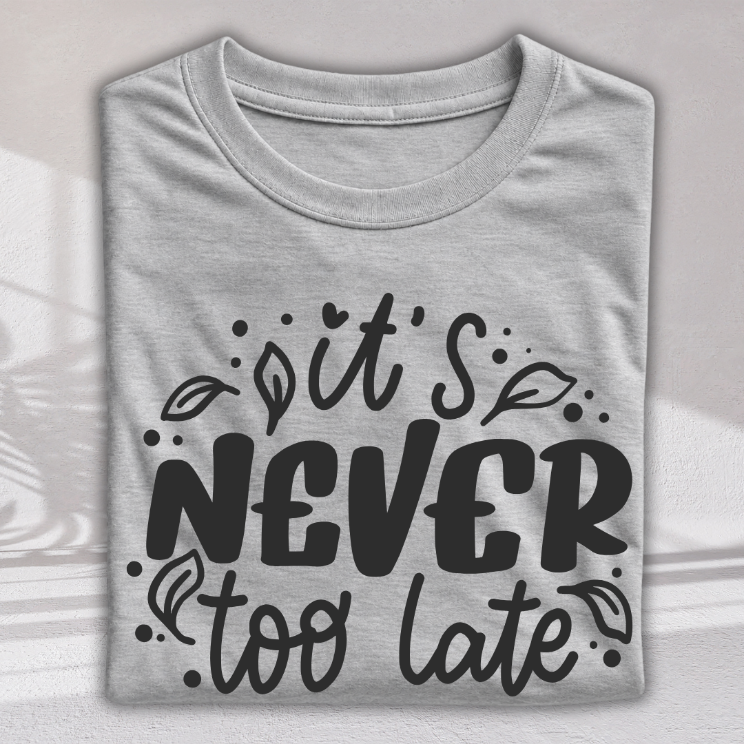 A folded light gray t-shirt with a black graphic print that reads 'it's NEVER too late' in a stylized hand-lettered font with small leaf and dot illustrations, displayed against a light gray background with soft shadow overlays.